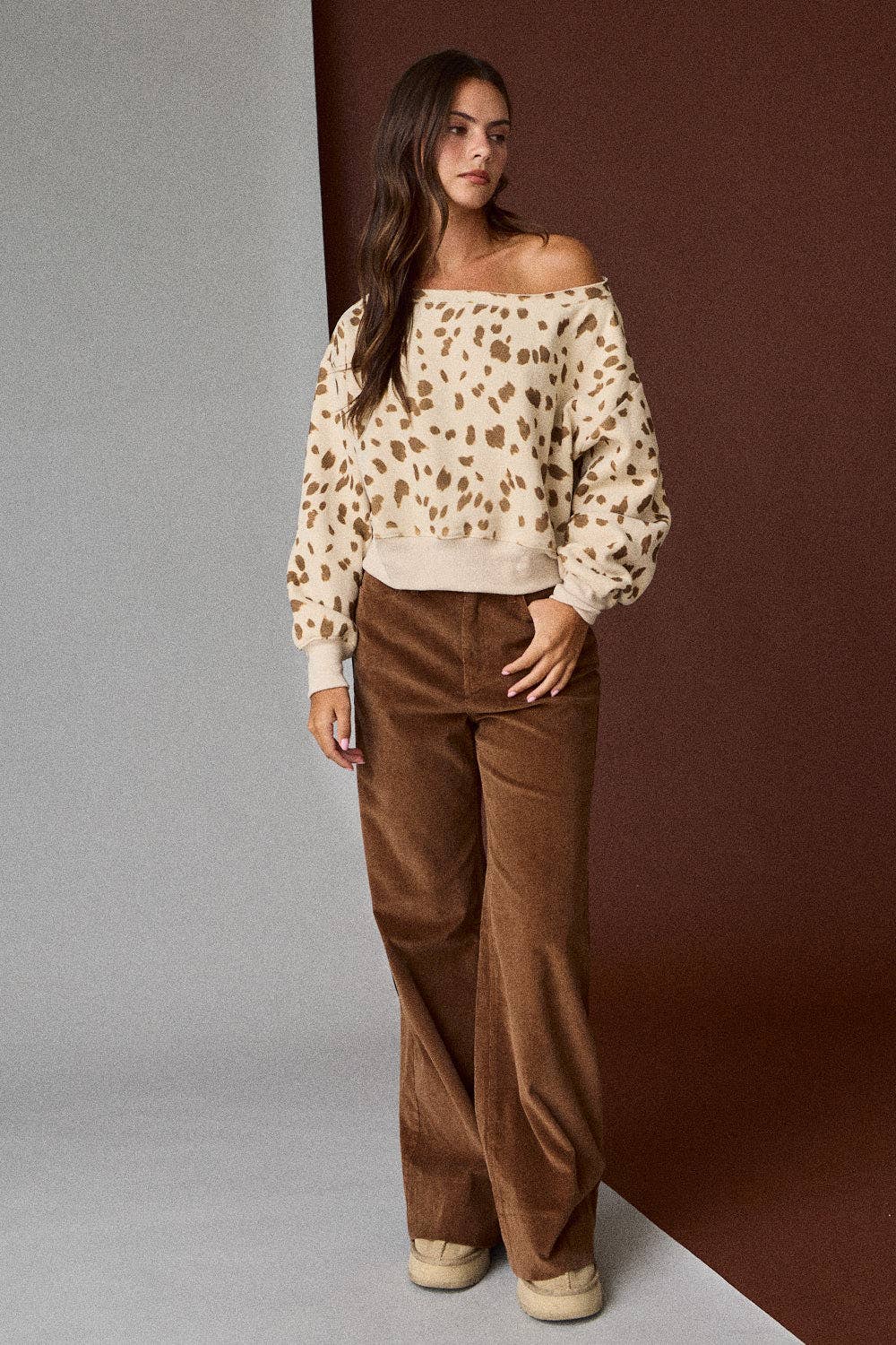 Woman wearing a leopard print off-shoulder top and brown pants against a gradient wall.