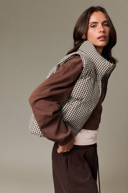 A model wearing a unique puffer vest with brown sleeves and a black-and-white houndstooth patterned body, styled with brown sweatpants against a solid gray studio background.