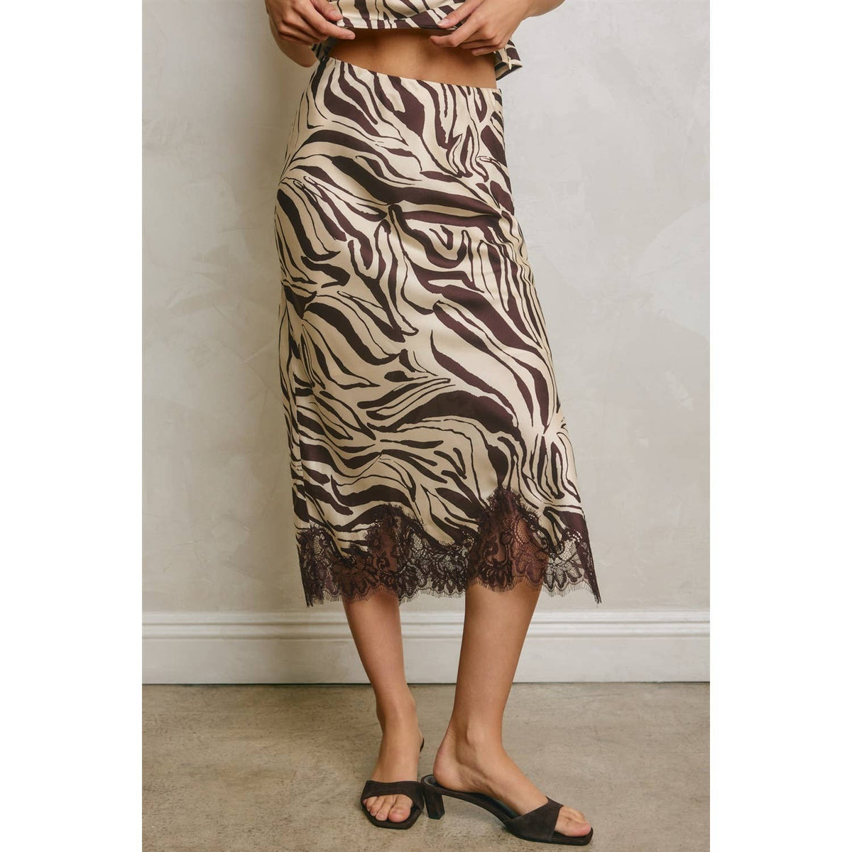 A model wearing a brown and beige zebra-print midi skirt with a lace hem, paired with black slide sandals, standing against a neutral wall.