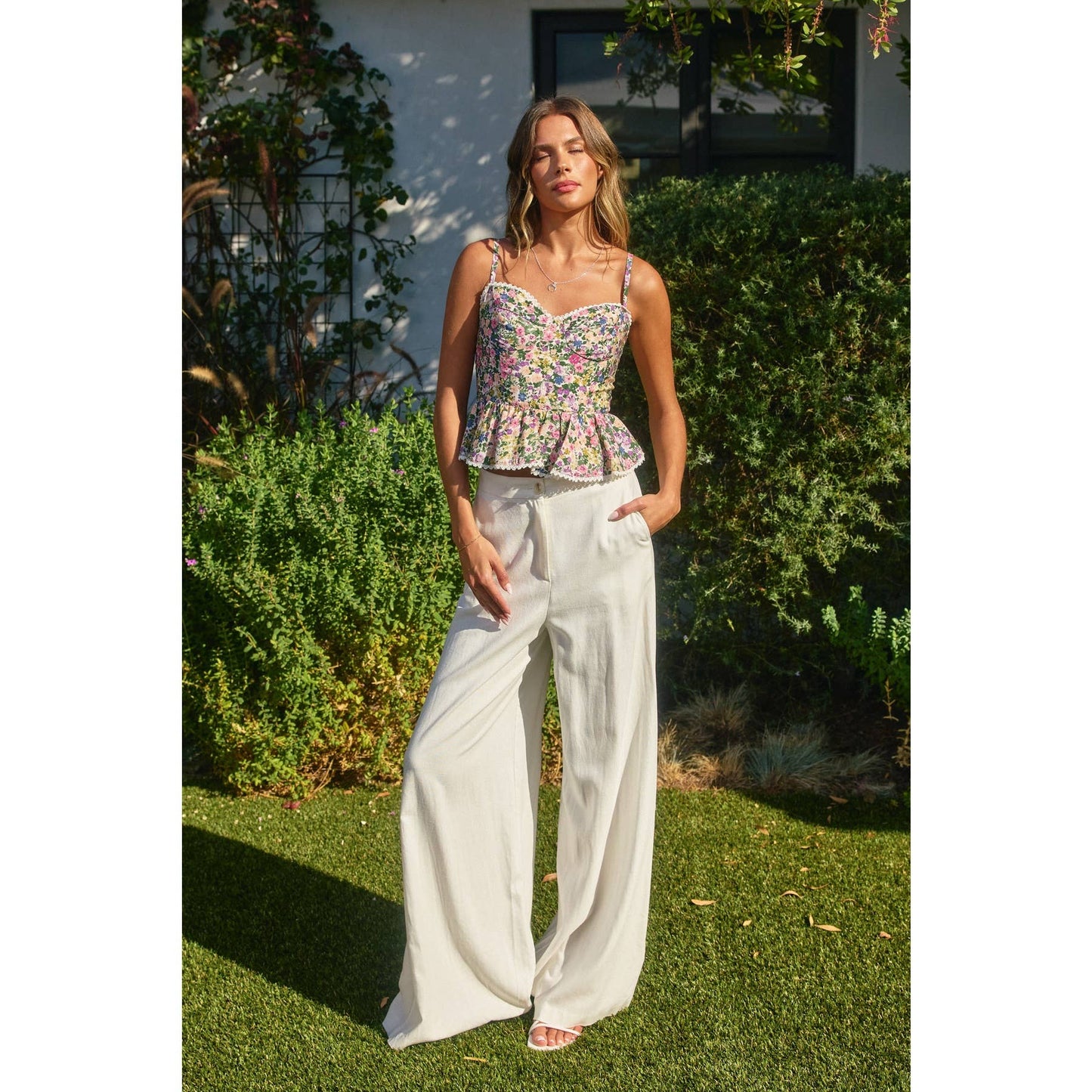 Woman wearing a floral top and white pants standing outdoors with greenery in the background