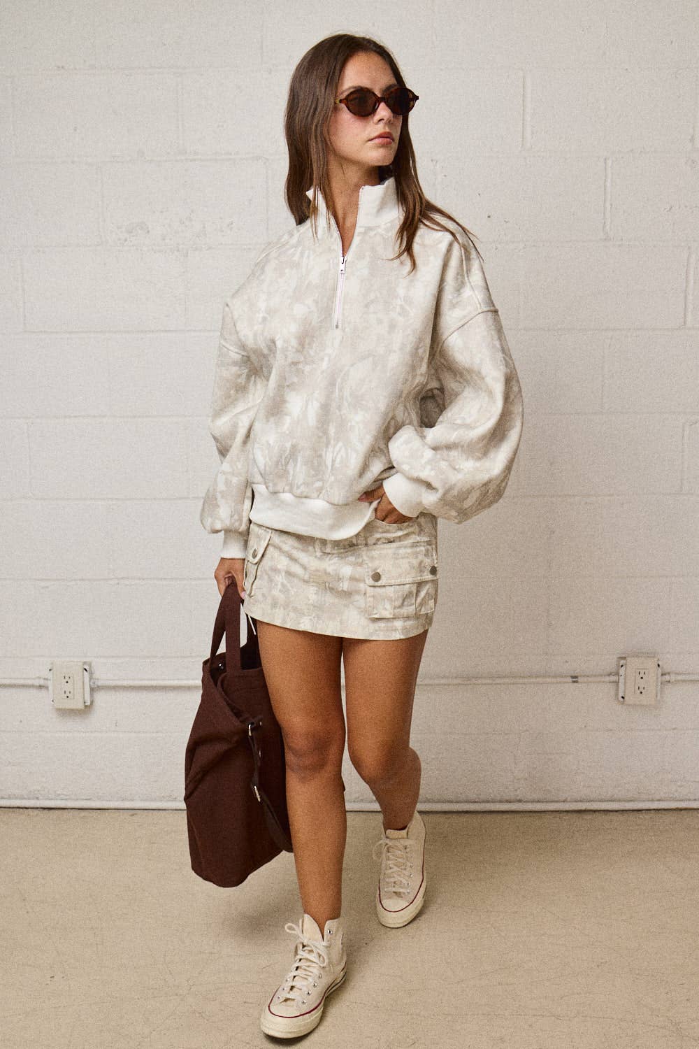 Woman wearing a white outfit with sunglasses and a brown bag against a white brick wall.