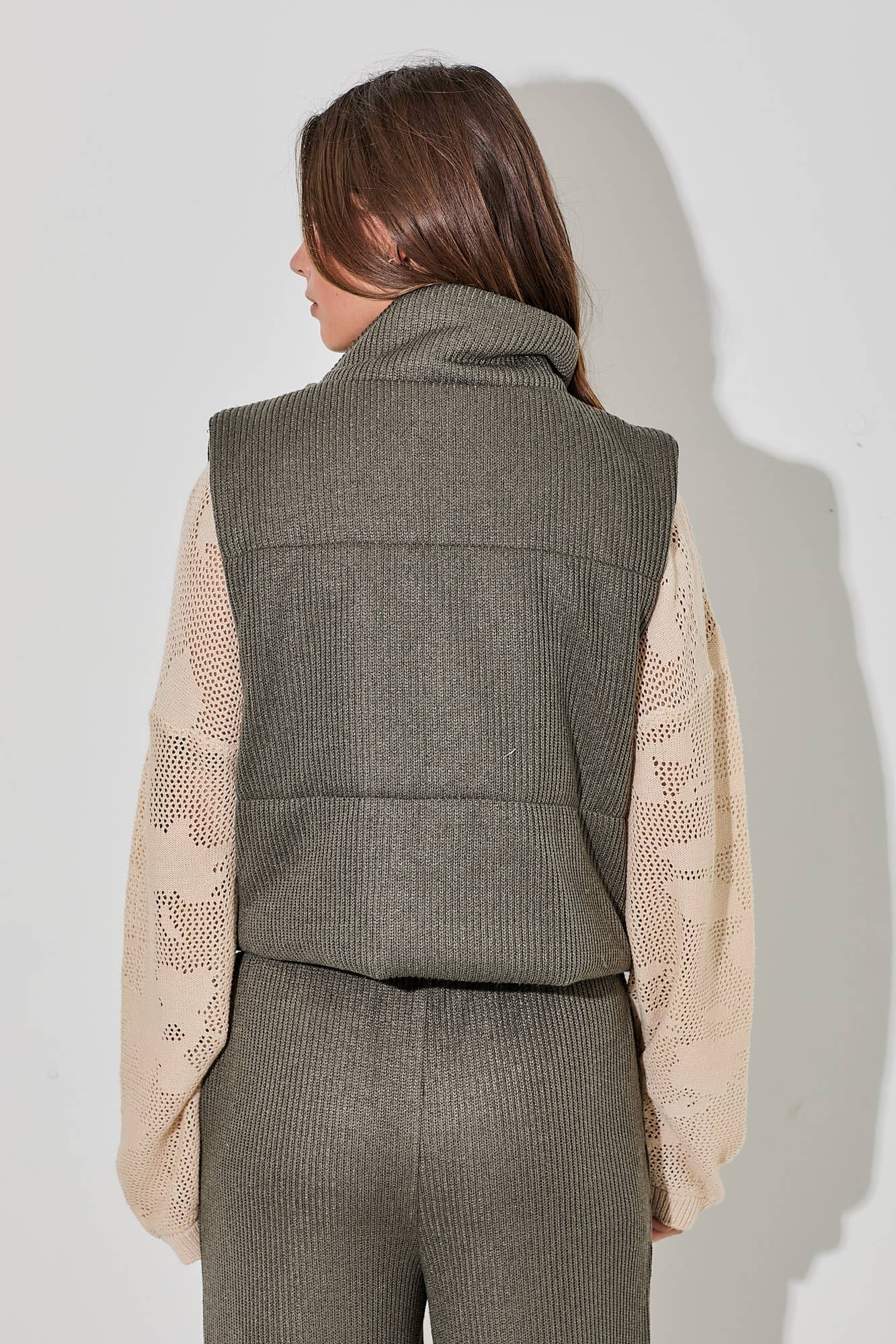 Person wearing a green vest over a beige sweater and gray pants on a white background