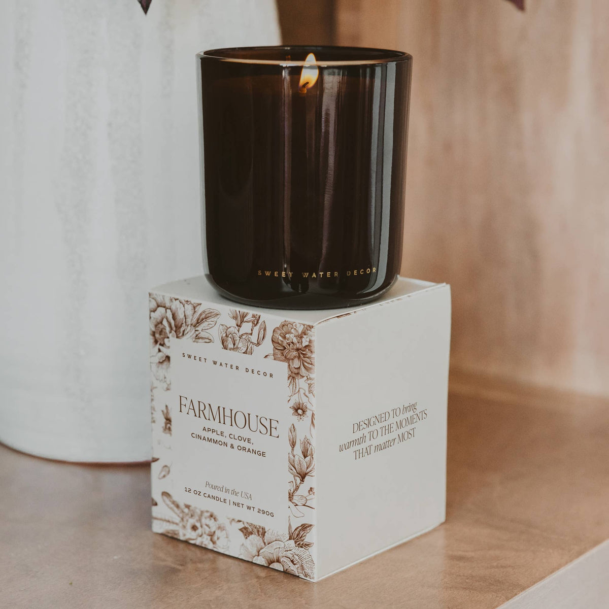 Black candle on a white box with floral design and 'Farmhouse' text, on a wooden surface.