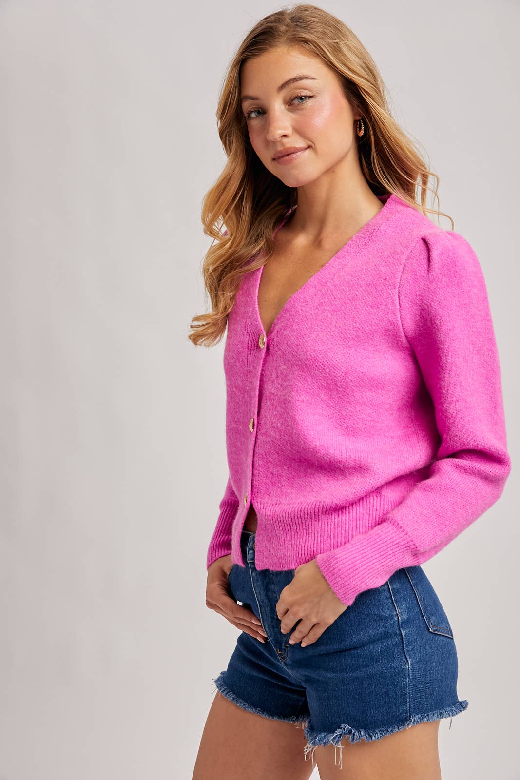 Woman wearing a bright pink cardigan and denim shorts on a plain background