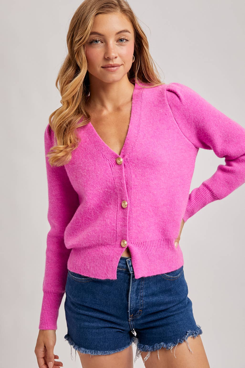 Woman wearing a bright pink cardigan with gold buttons on a plain background