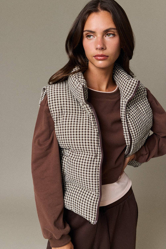 A woman modeling a sleeveless puffer vest with a black and white houndstooth pattern over brown long-sleeve sweatshirt sleeves, paired with a brown top and dark brown pants, against a solid taupe background.