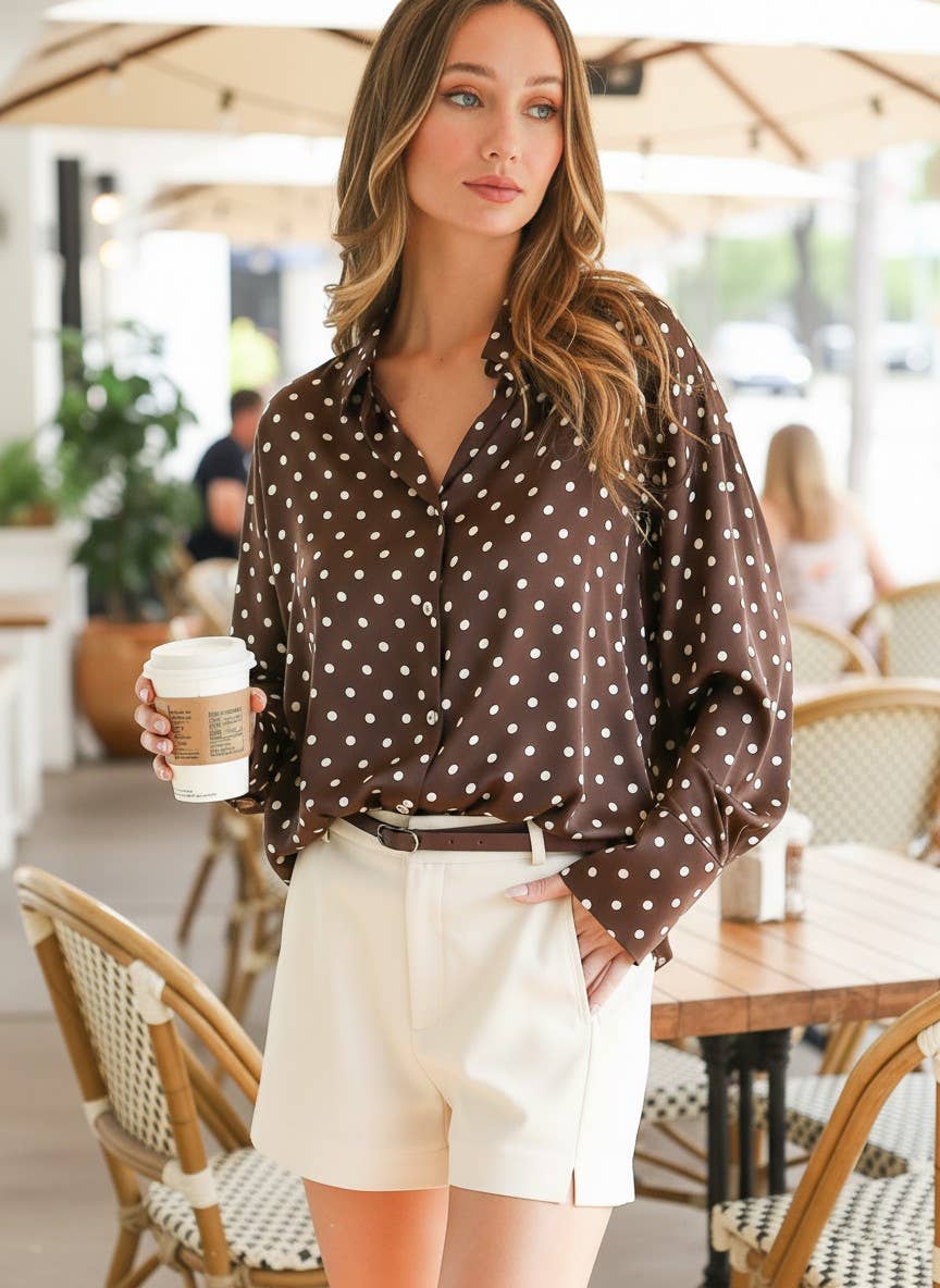 Woman in a polka dot blouse and shorts holding a coffee cup in an outdoor cafe setting.