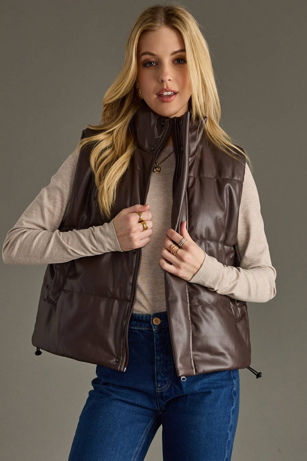 Woman wearing a brown vest over a beige sweater and blue jeans on a gray background