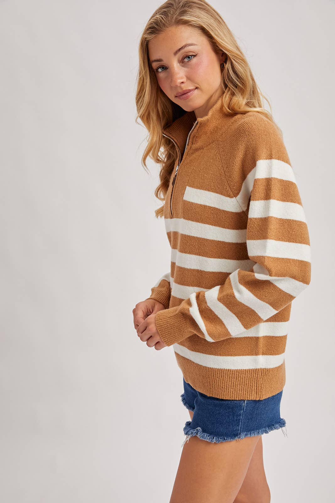 Woman wearing a brown and white striped sweater with denim shorts on a plain background