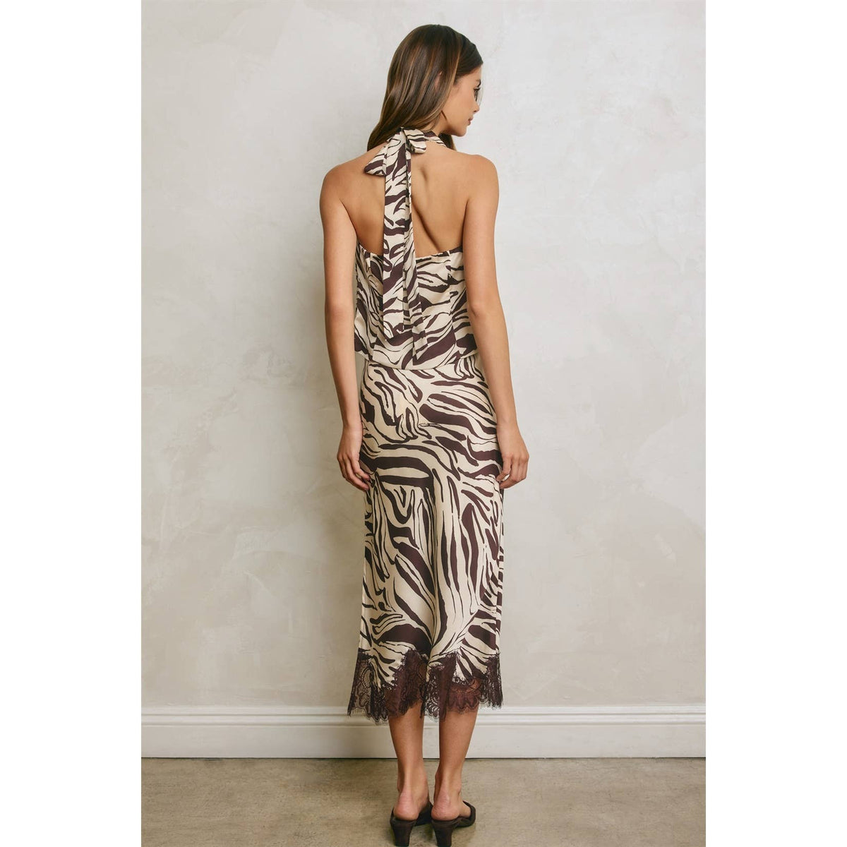A woman stands with her back to the camera wearing a sleeveless halter-neck midi dress with a brown and beige zebra print and a ruffled lace hem, paired with black heels.