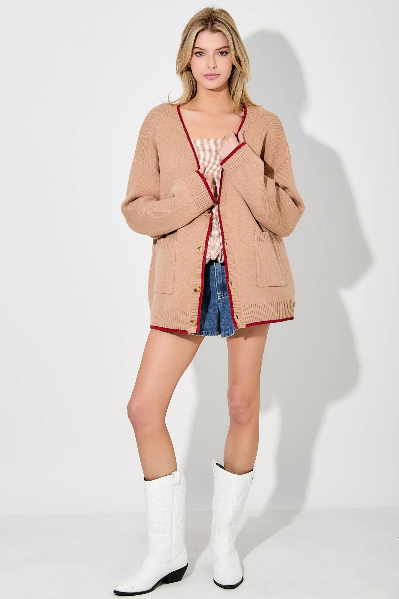 Woman wearing a beige cardigan with red trim, denim shorts, and white boots on a white background