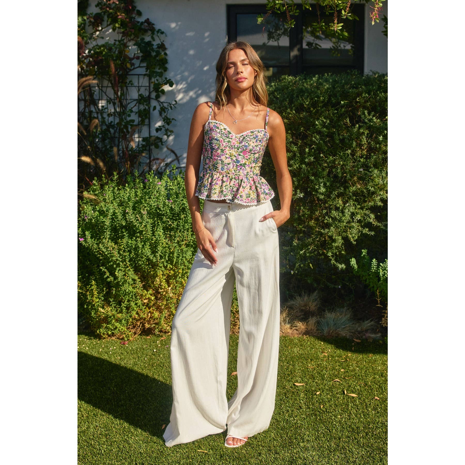 Woman wearing a floral top and white pants standing outdoors with greenery in the background