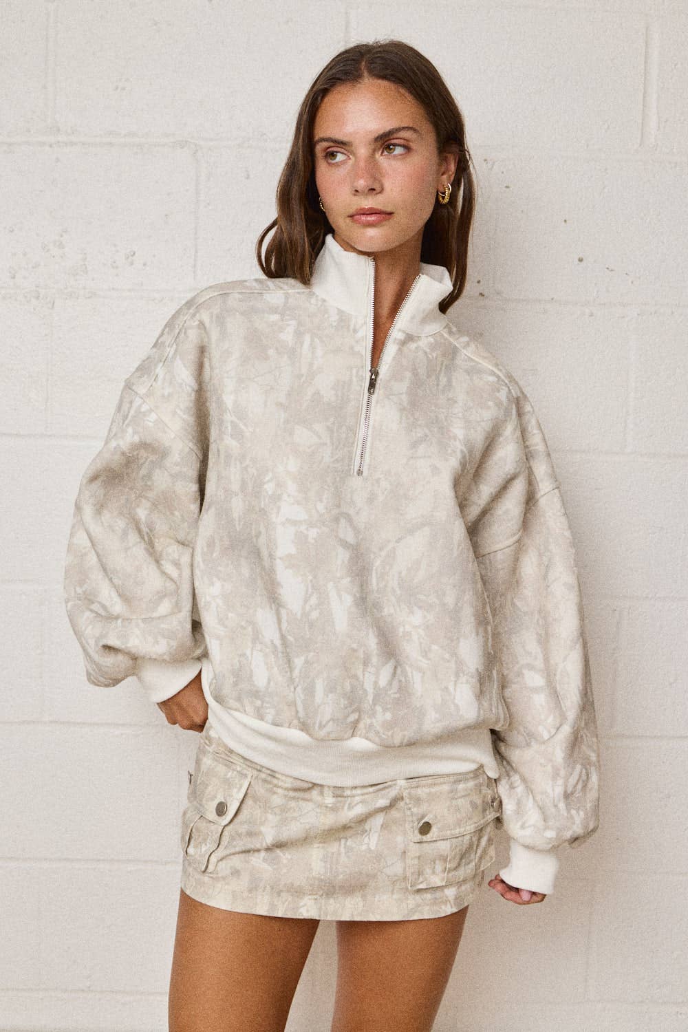 Woman wearing a beige fleece pullover and shorts against a white brick wall.