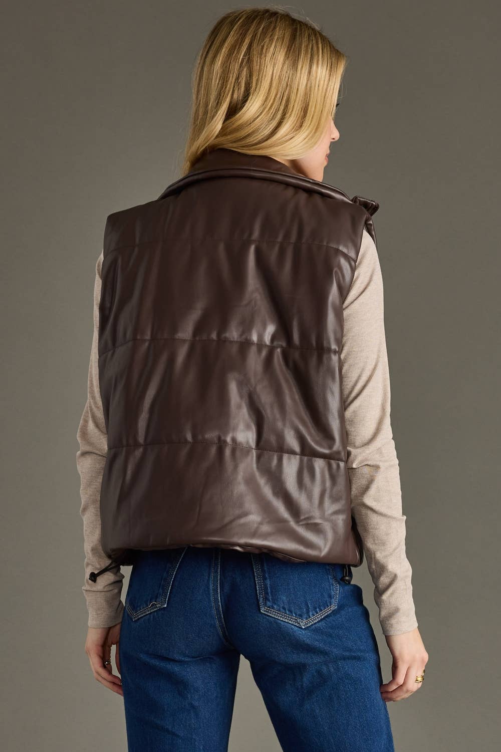 Person wearing a brown leather vest over a beige shirt and blue jeans on a gray background