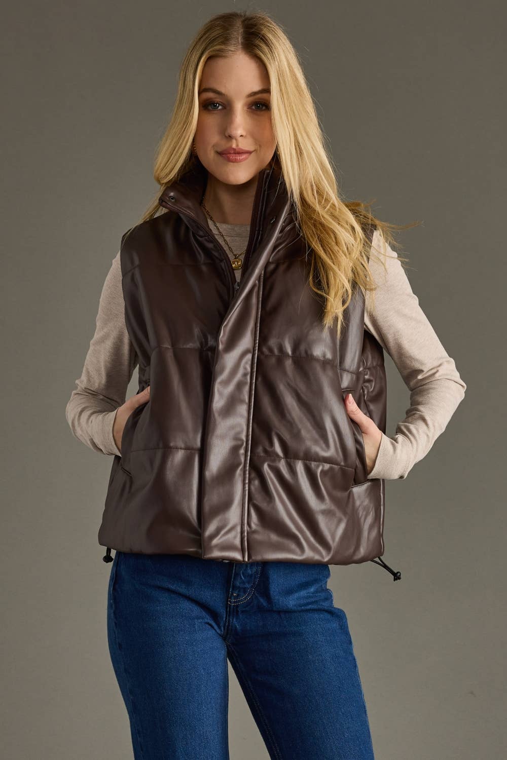 Woman wearing a brown puffer vest over a light gray shirt and blue jeans on a gray background