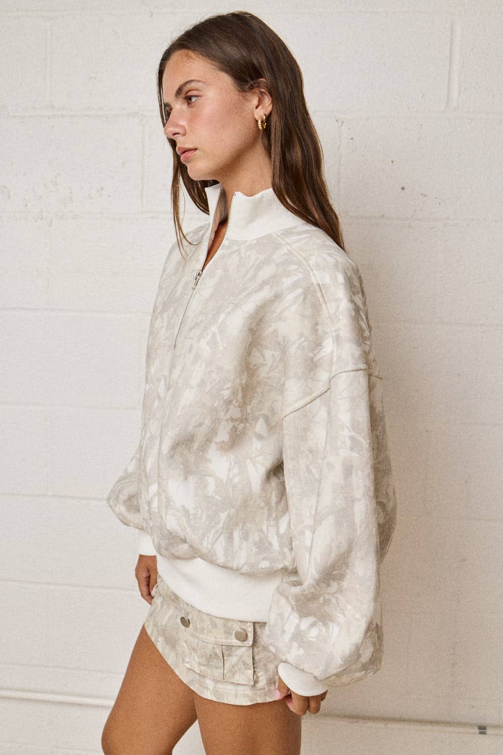 Woman wearing a white tie-dye jacket and shorts against a white brick wall.