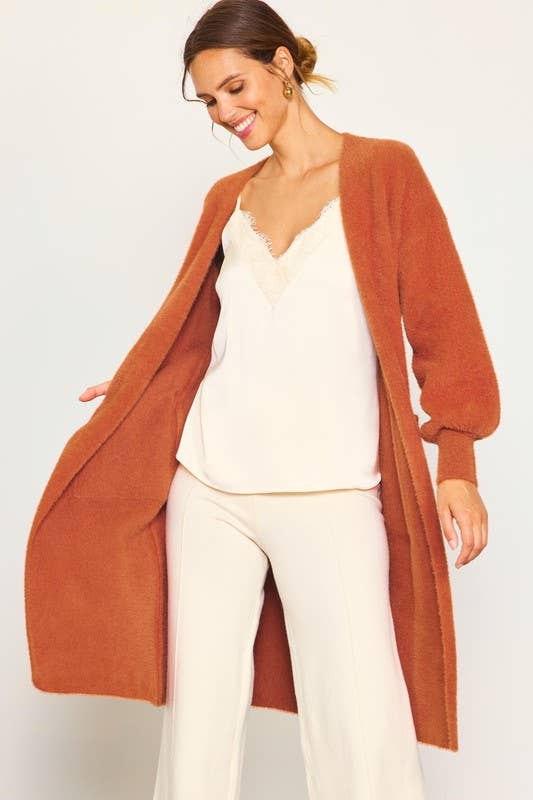 Woman wearing a long rust-colored cardigan over a white top and pants on a light gray background