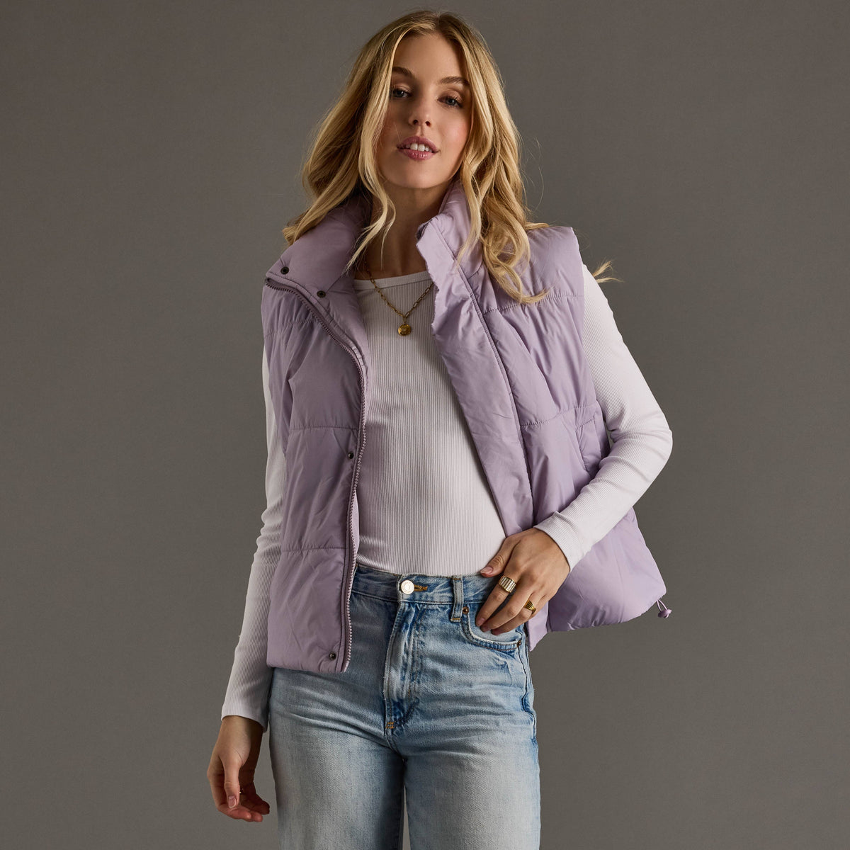 Woman wearing a light purple puffer vest over a white shirt and blue jeans on a gray background