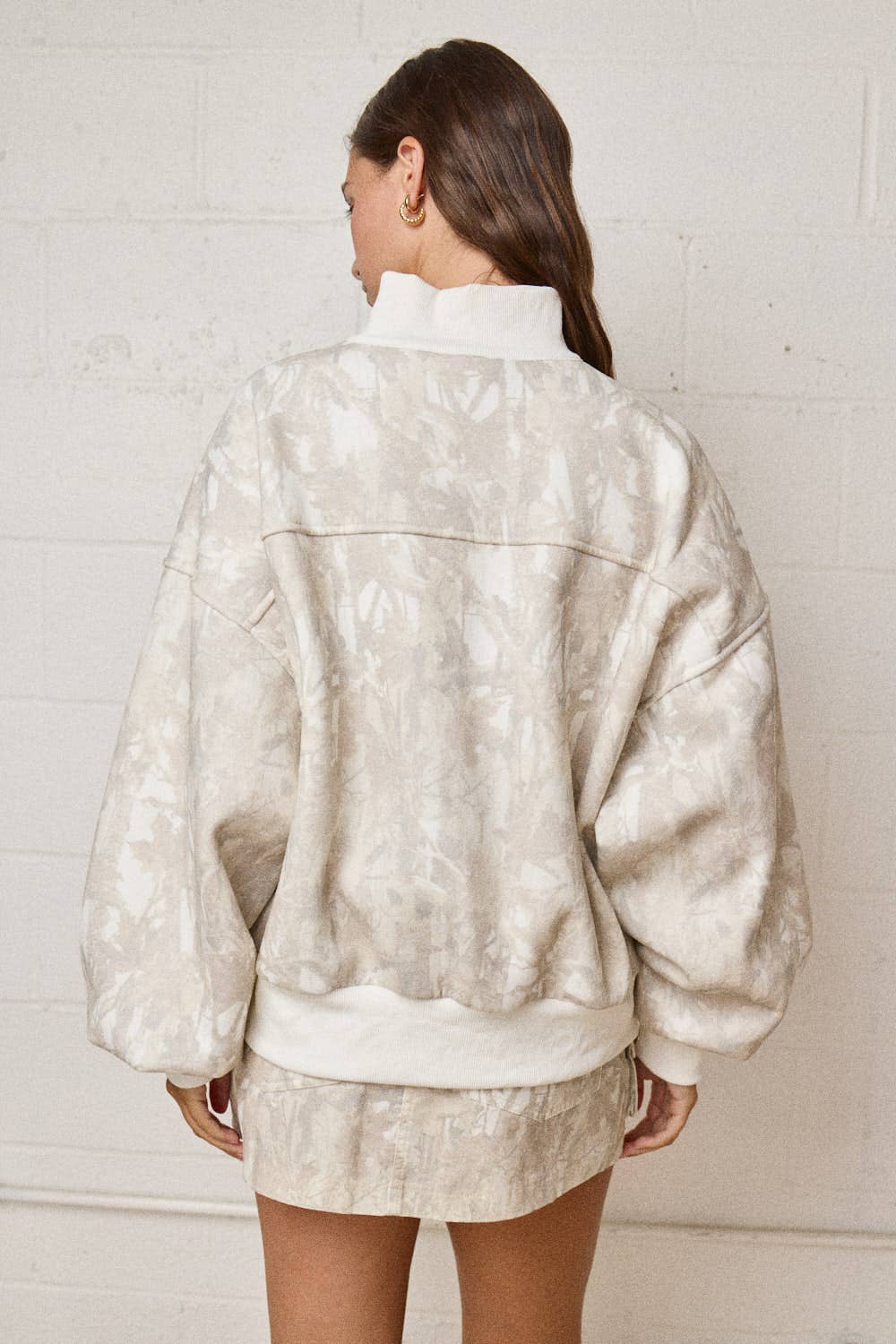 Person wearing a beige patterned jacket and shorts against a white brick wall.