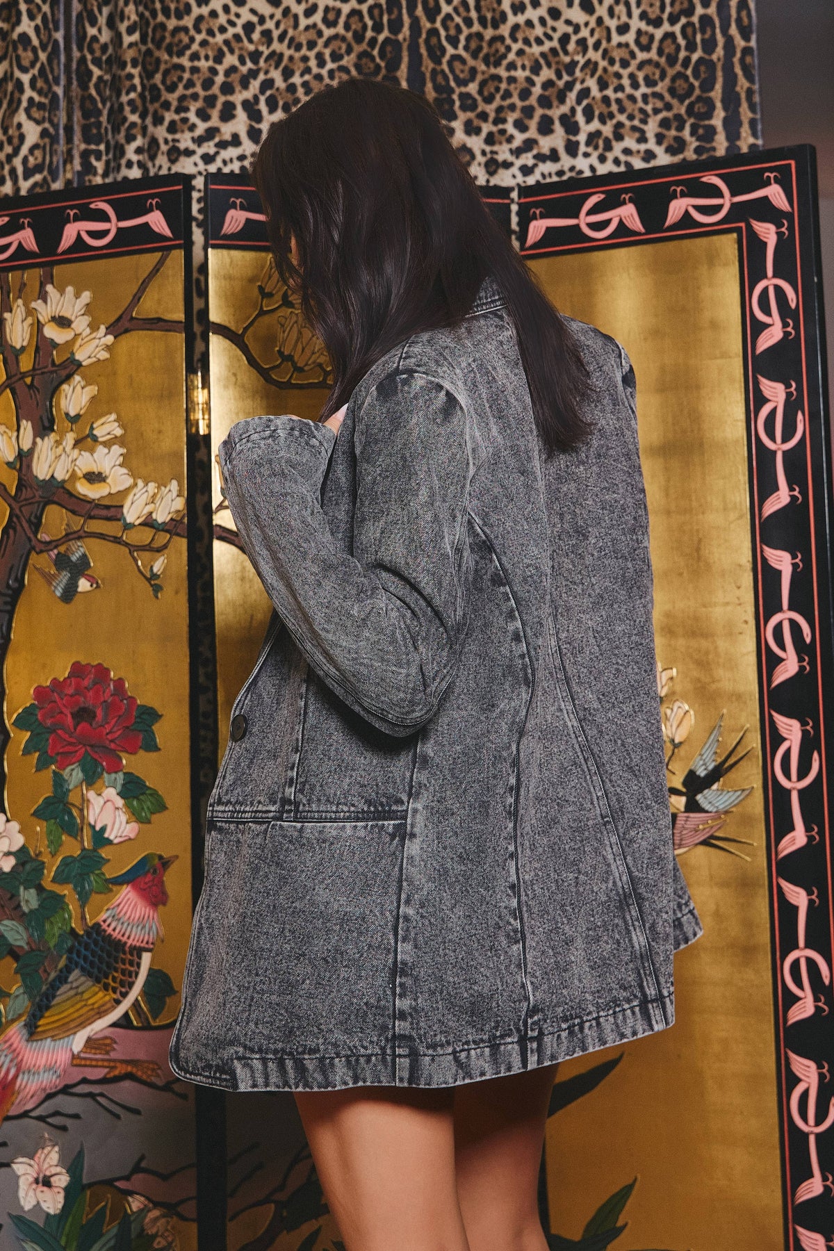 A woman wearing a faded gray denim jacket stands in front of a decorative gold folding screen with floral and bird motifs.
