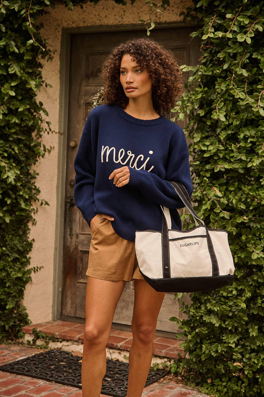 A woman stands outdoors in front of a door framed by green ivy, wearing a navy blue knit sweater with the word 'merci' in white script across the chest, tan shorts, and carrying a large cream tote bag with black trim and the brand name 'MERCI" visible on the front.