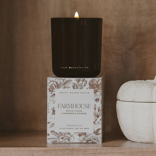 A lit black candle in a matte black vessel sits on top of its floral-patterned box, which reads 'Sweet Water Decor' and 'Farmhouse'. The scene is styled on a wooden surface with a warm brown textured background and a white ceramic object partially visible on the right.
