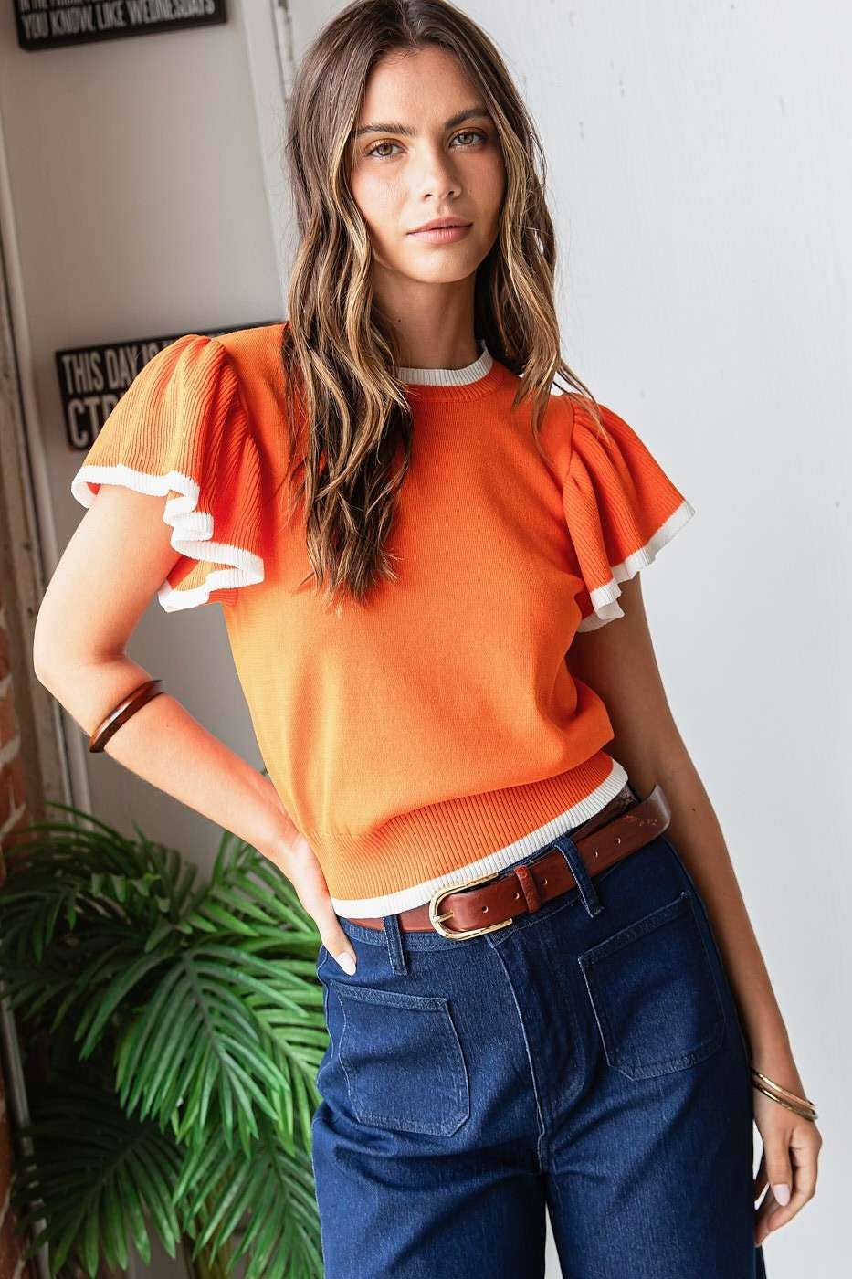 Woman wearing an orange top with white trim and blue pants indoors.