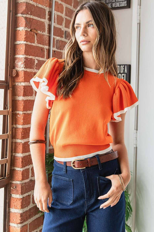 Woman wearing an orange top with ruffled sleeves and blue jeans standing against a brick wall.