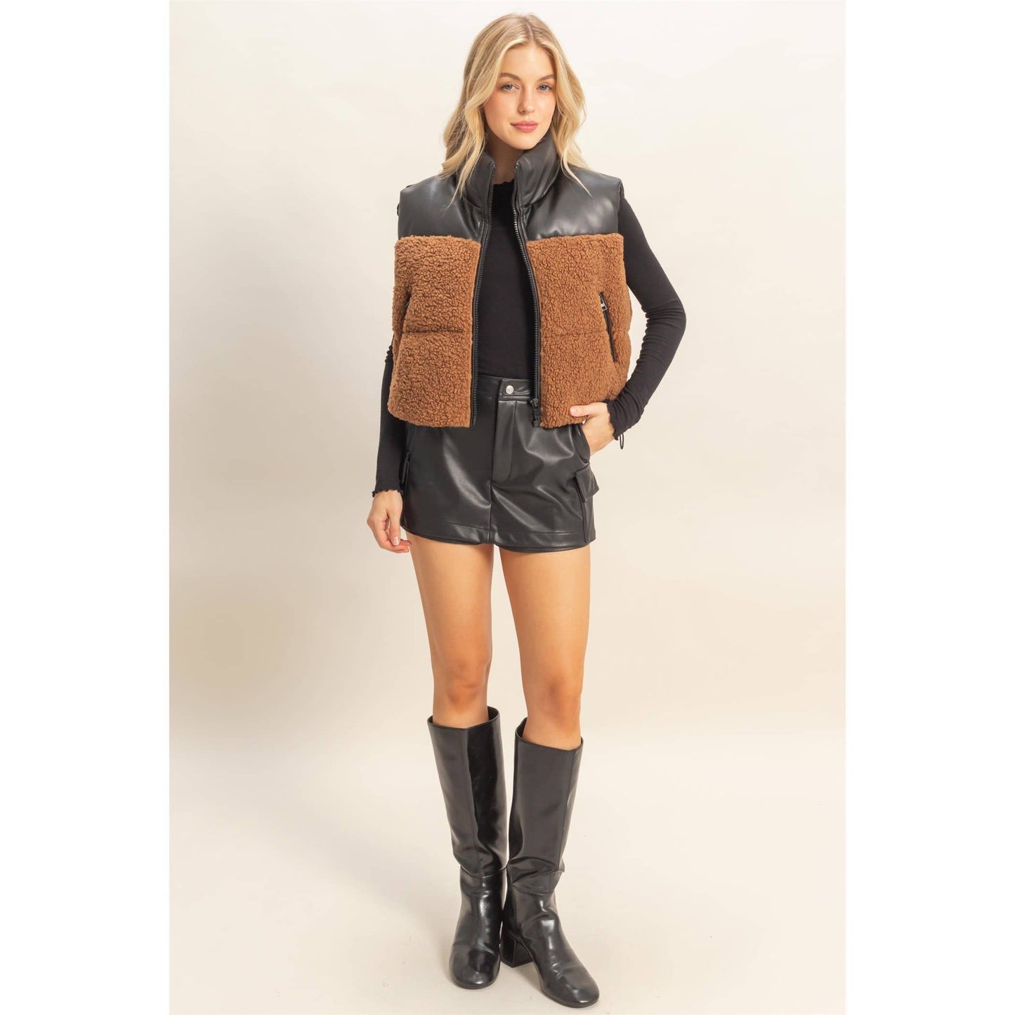 Woman wearing a brown and black quilted vest with black leather shorts and boots on a white background