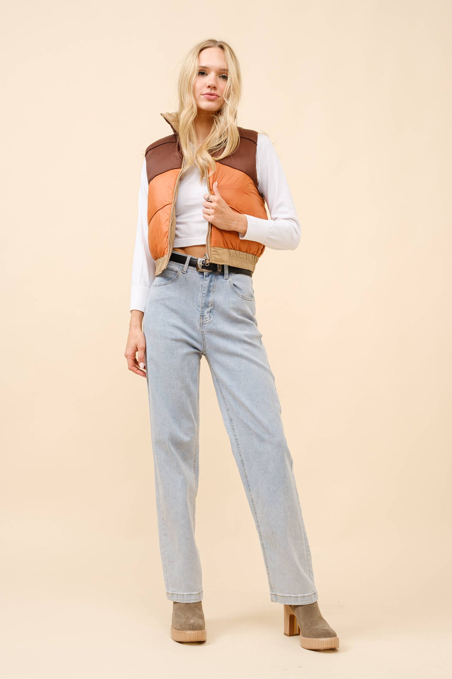 Woman wearing a two-tone vest, white shirt, light blue jeans, and brown boots on a beige background
