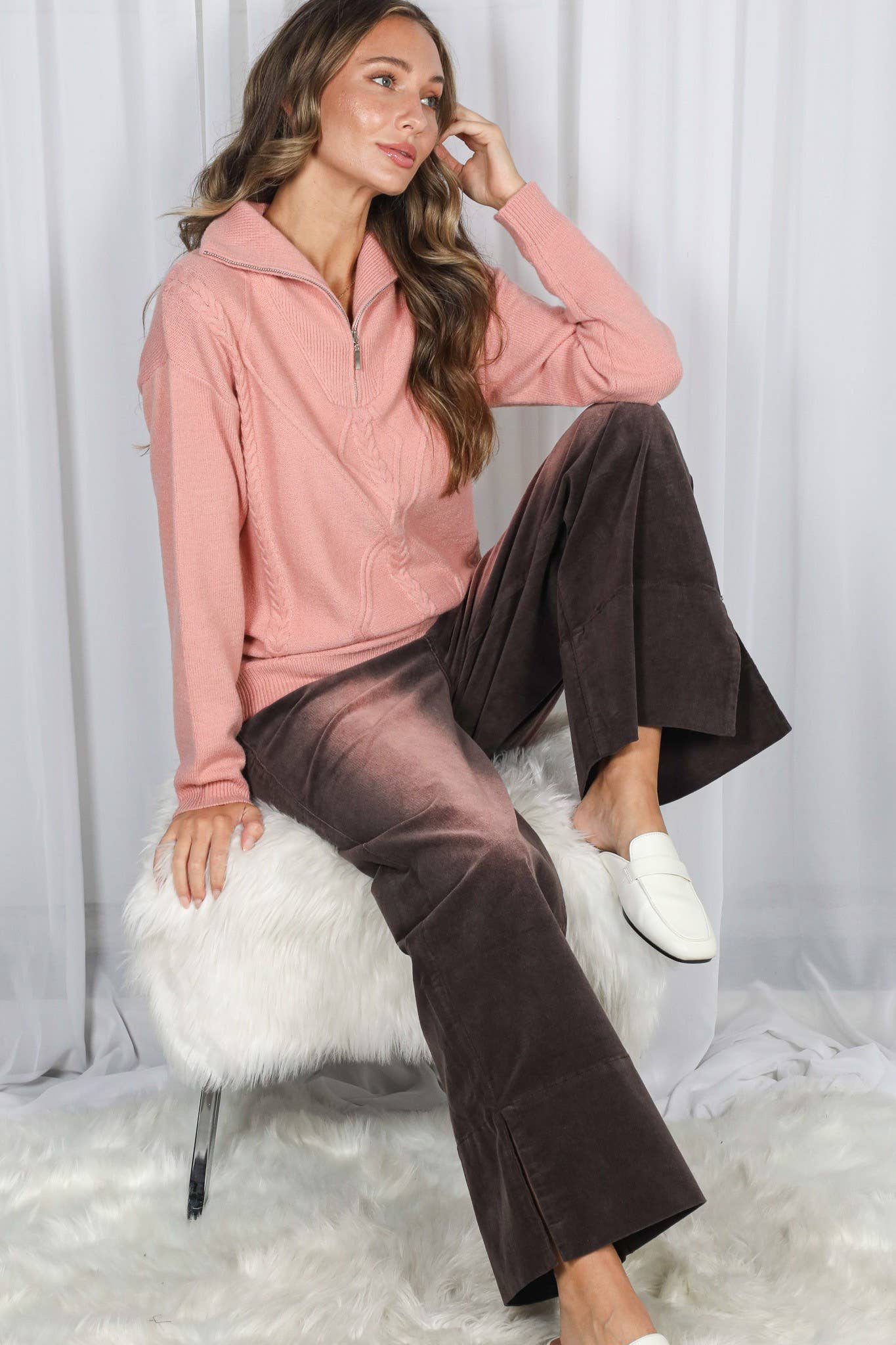 Woman sitting on a white stool wearing a pink hoodie and brown pants against a white curtain background.