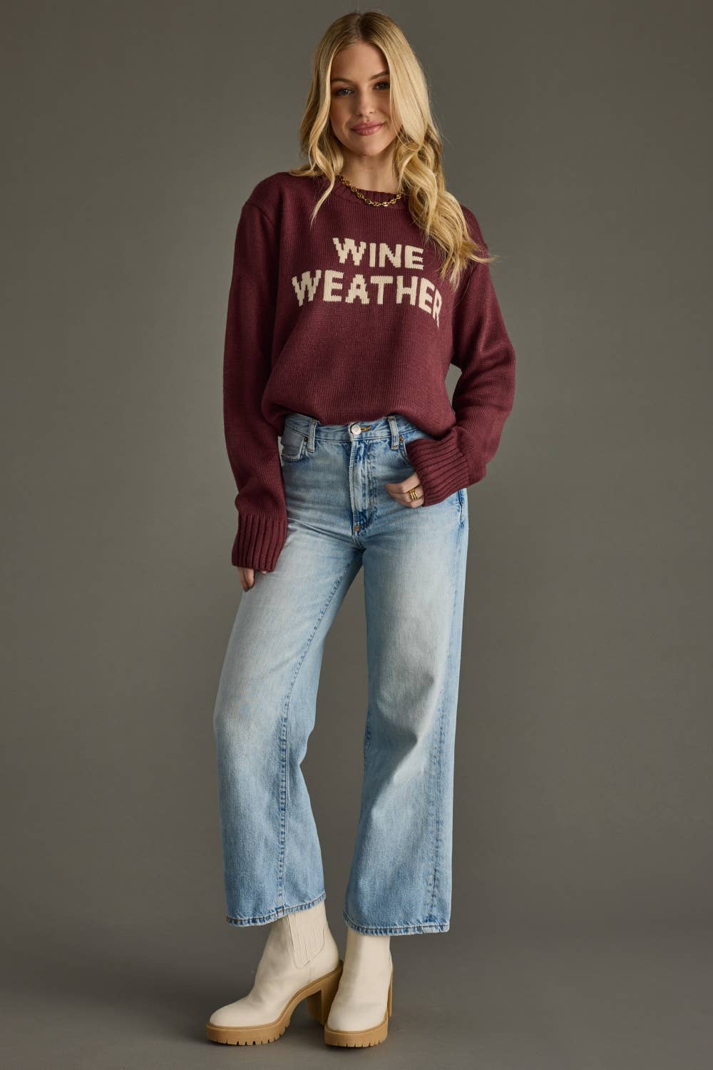 Woman wearing a maroon sweater with 'Wine Weather' text and blue jeans on a gray background